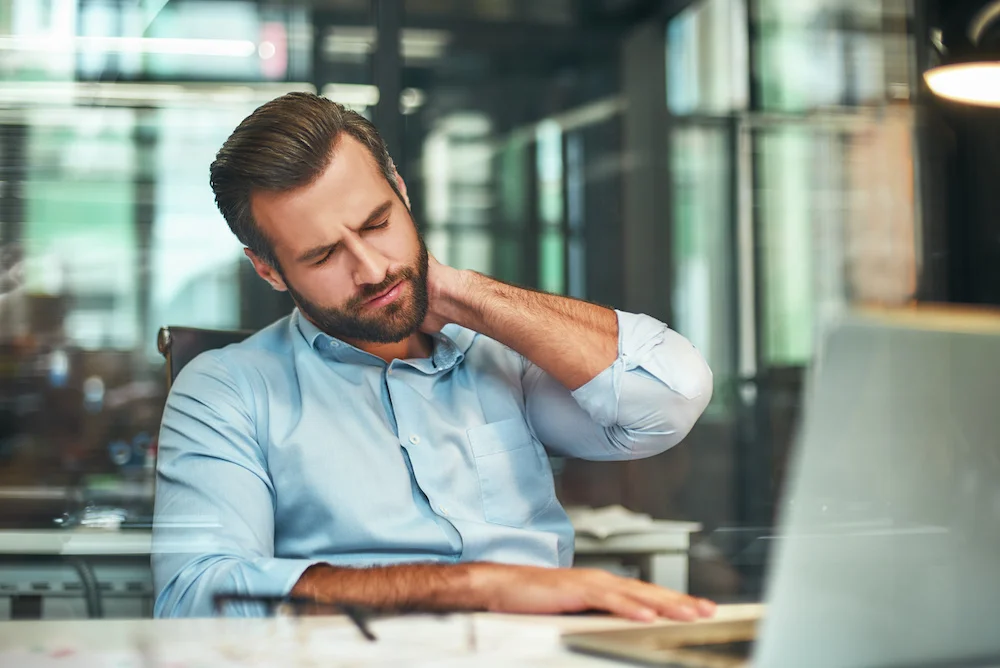 A man holds the side of his neck with his left hand while sitting at a desk