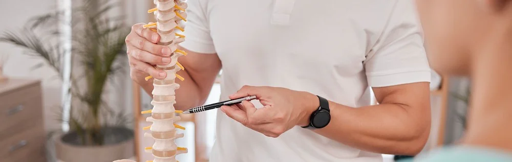 A doctor presses a pen into a model of a spine while speaking to a patient