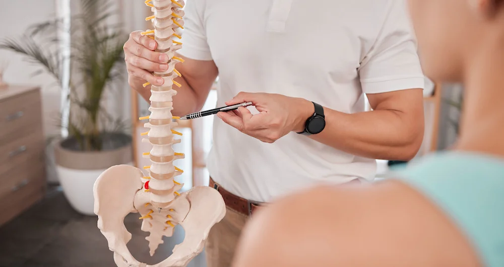 A doctor presses a pen into a model of a spine while speaking to a patient