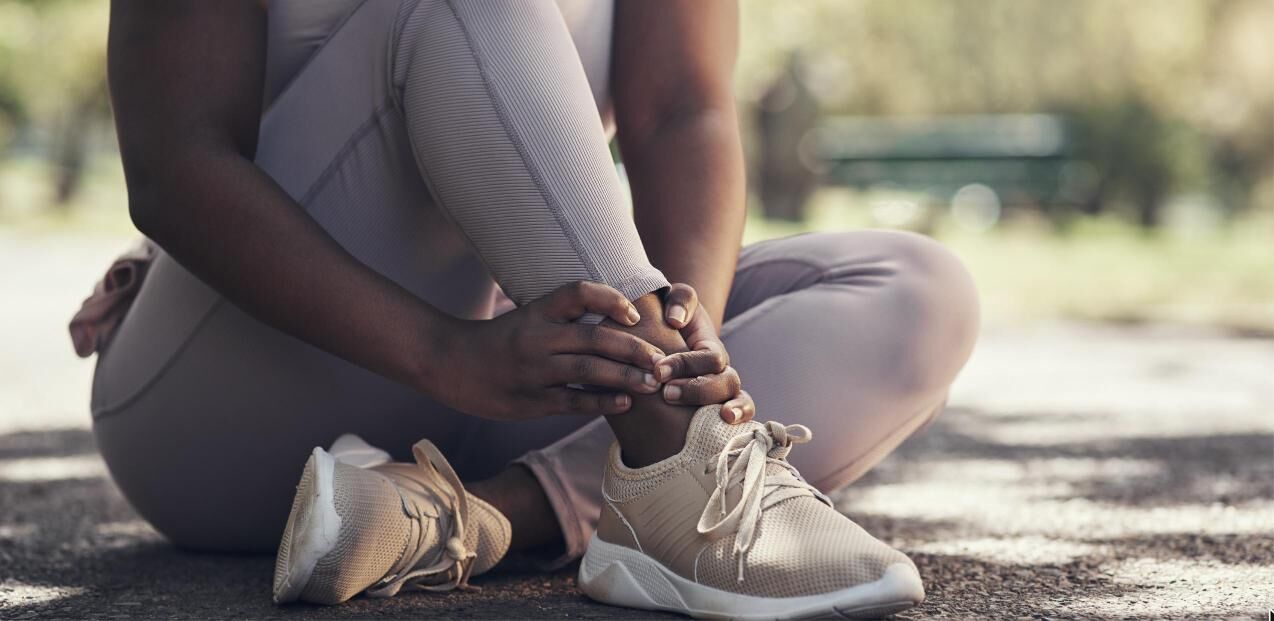 A woman sits on a gravel trail while gripping her right ankle.